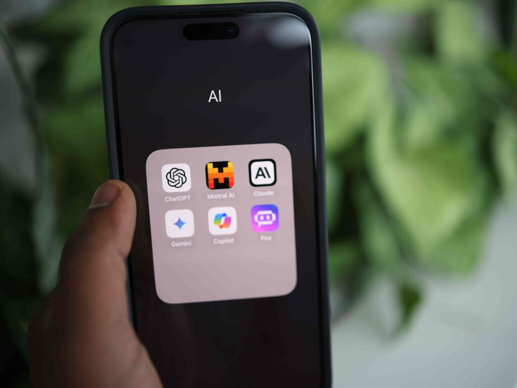 A close-up of a person holding a smartphone showing an open folder named "AI" with icons for ChatGPT, Mistral AI, Claude, Gemini, Copilot, and Poe, set against a green plant background.