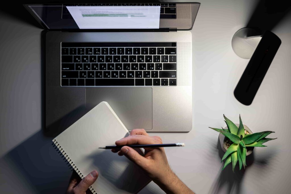 Top-down view of a person holding a pencil and writing in a spiral notebook, positioned below an open laptop under a desk lamp with a small succulent plant nearby.
