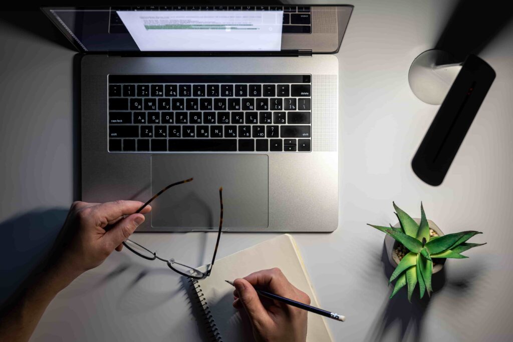 Top-down view of a person holding eyeglasses and writing in a spiral notebook beside an open laptop illuminated by a desk lamp at night.