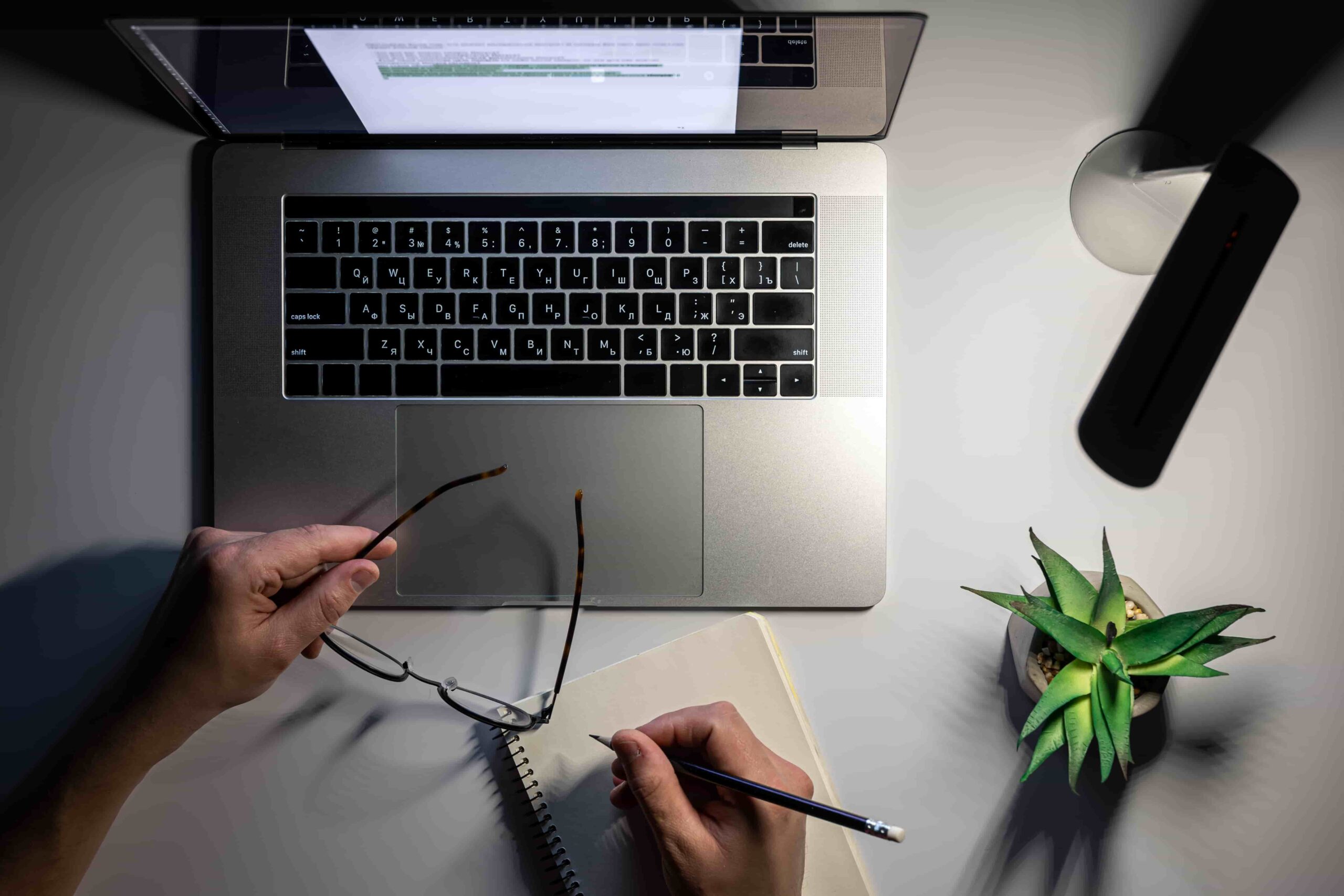 Top-down view of a person holding eyeglasses and writing in a spiral notebook beside an open laptop illuminated by a desk lamp at night.
