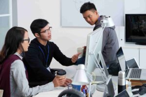 A group of three young professionals and software developers having a collaborative discussion around a desktop computer in a modern office environment.