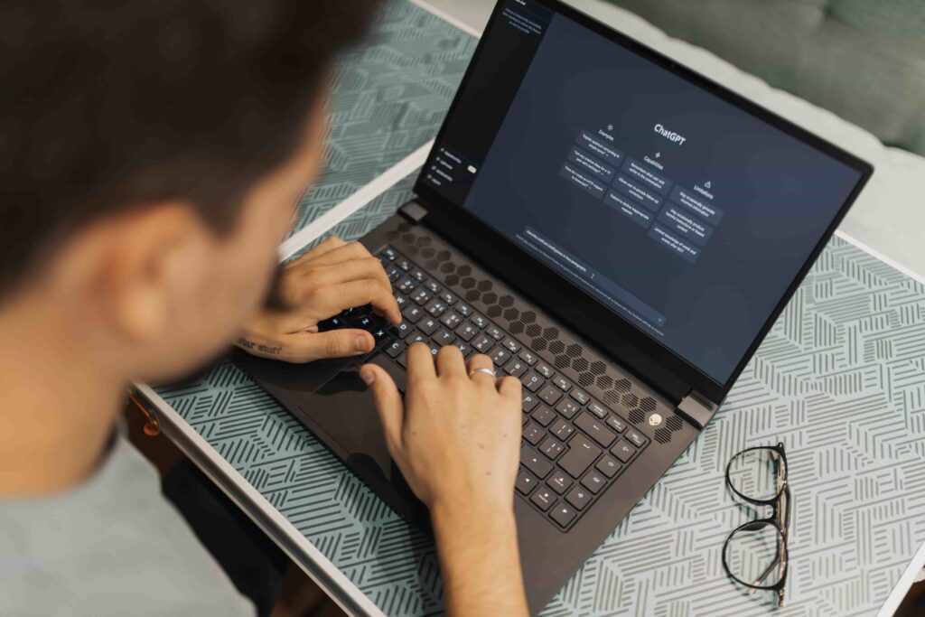 An over-the-shoulder shot of a person typing on a laptop keyboard with the ChatGPT dark interface open on screen, placed on a patterned tray next to a pair of glasses.