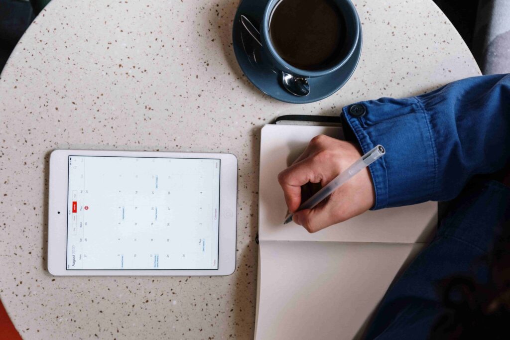 Top-down view of a person wearing a denim jacket writing in a notebook next to a digital tablet displaying a calendar app and a cup of black coffee on a round table.