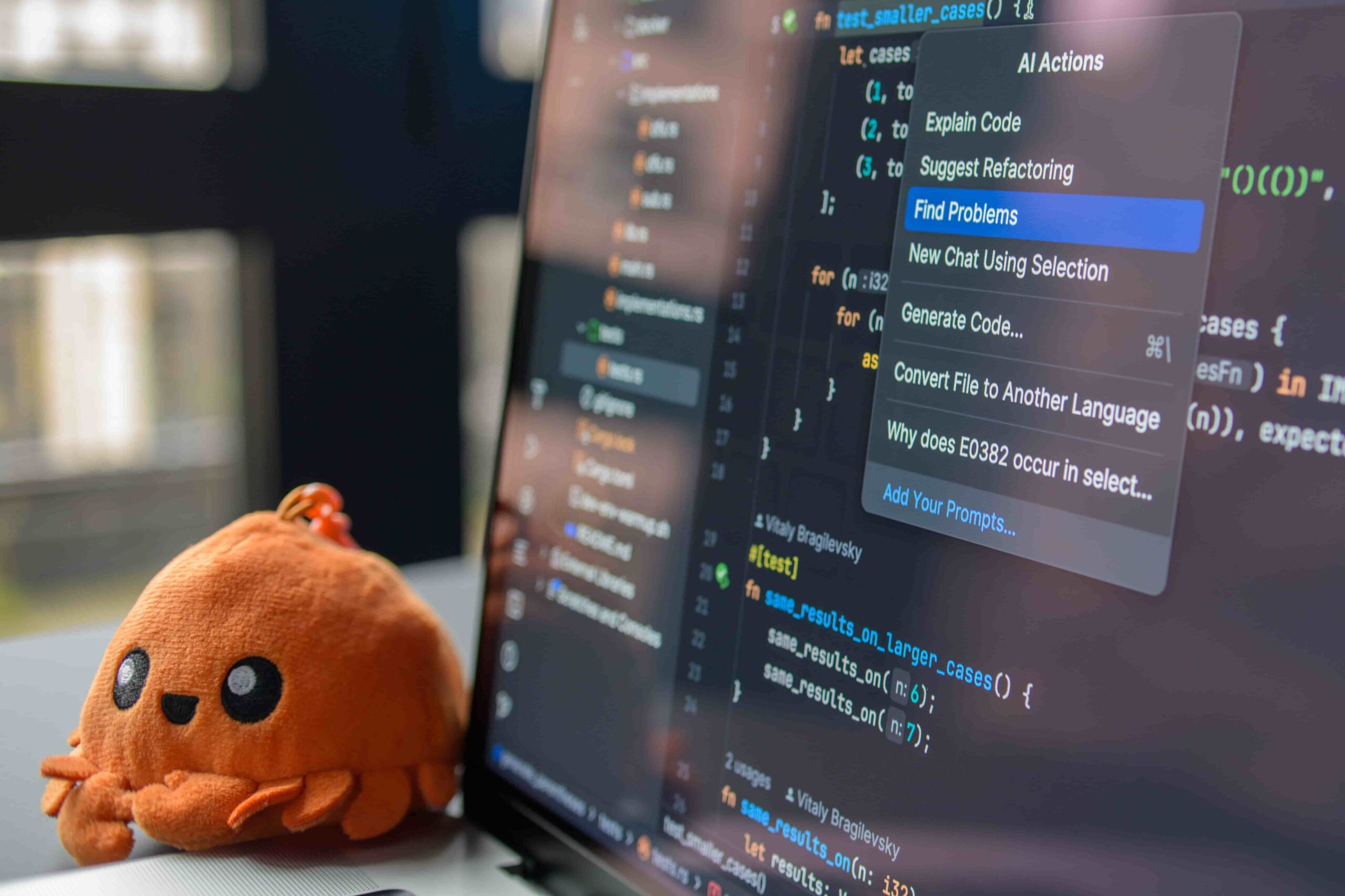 A close-up of a laptop screen showing code in a code editor with an AI Actions menu open, displaying options like Explain Code and Find Problems, with an orange plush toy sitting next to the computer.