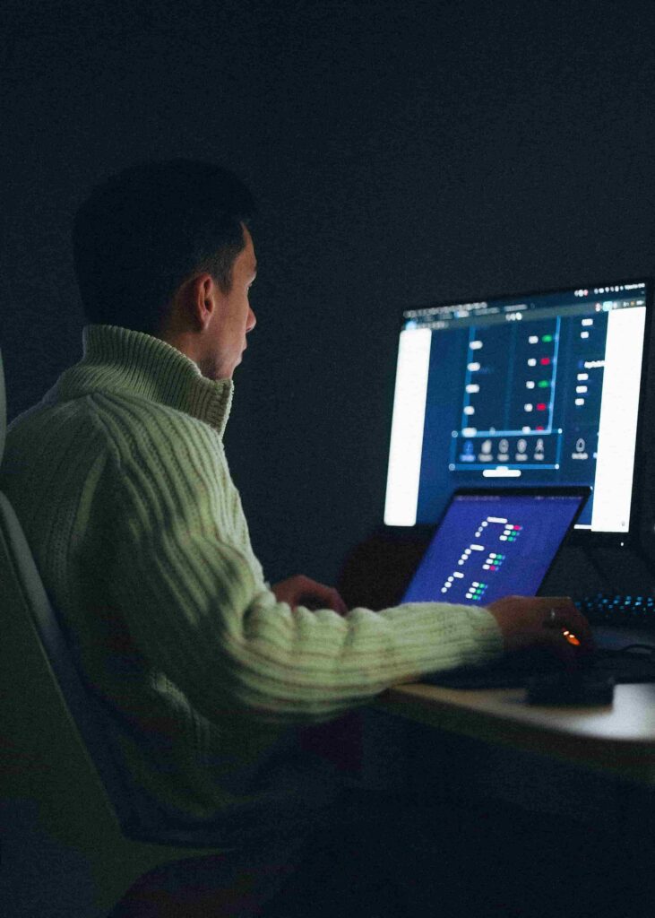 A man wearing a high-neck sweater working at night in a dimly lit room, analyzing charts and data across a laptop and a large desktop monitor.
