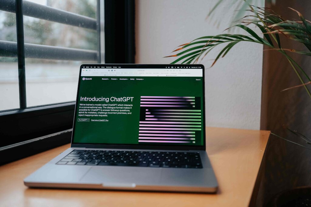 A laptop open on a wooden desk near a window displaying OpenAI's "Introducing ChatGPT" webpage with a green houseplant on the side.