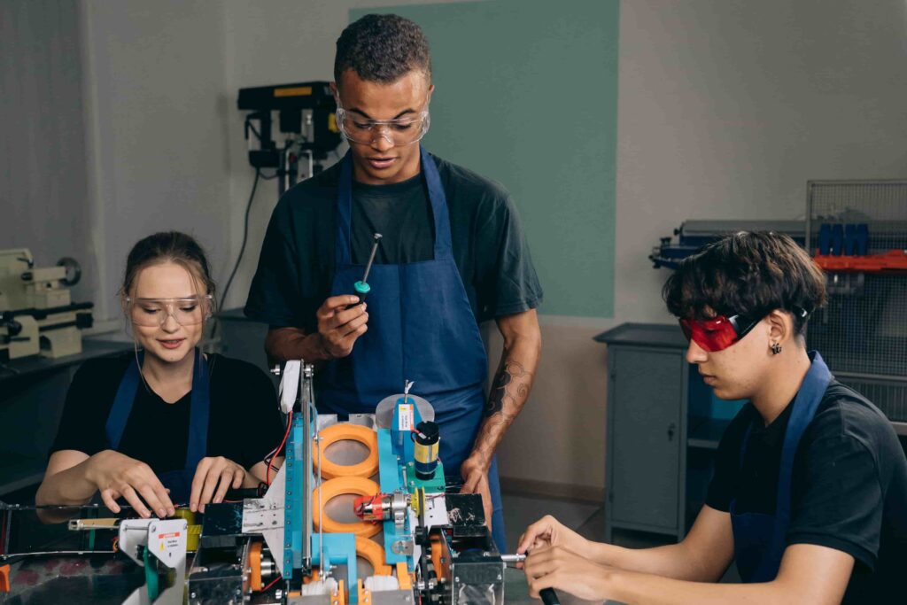 Three young engineering students wearing protective safety glasses working together on a complex robotics project inside a technical workshop.