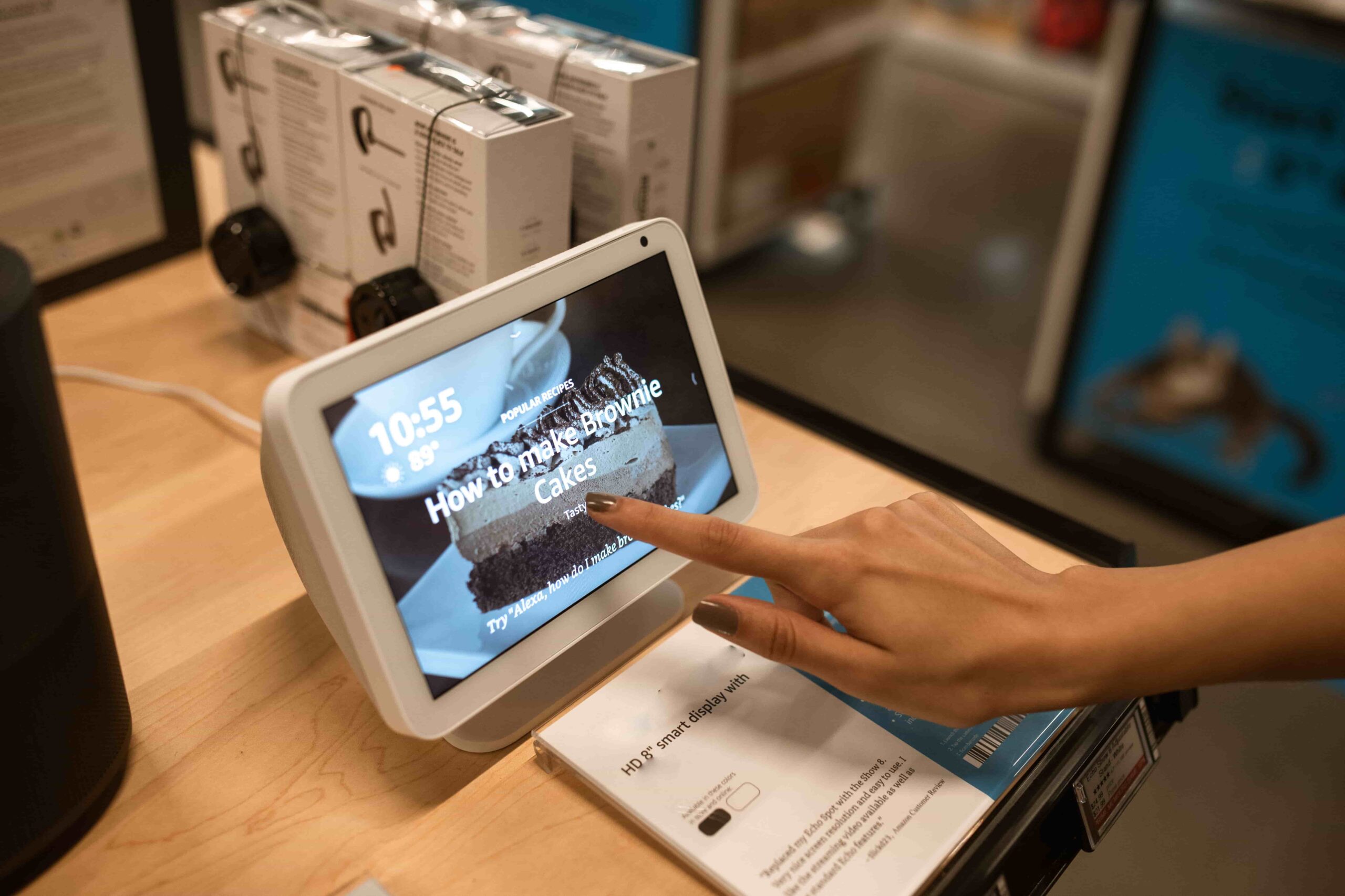 A person's hand touching the screen of a smart display showing a brownie cake recipe prompt inside a retail store.